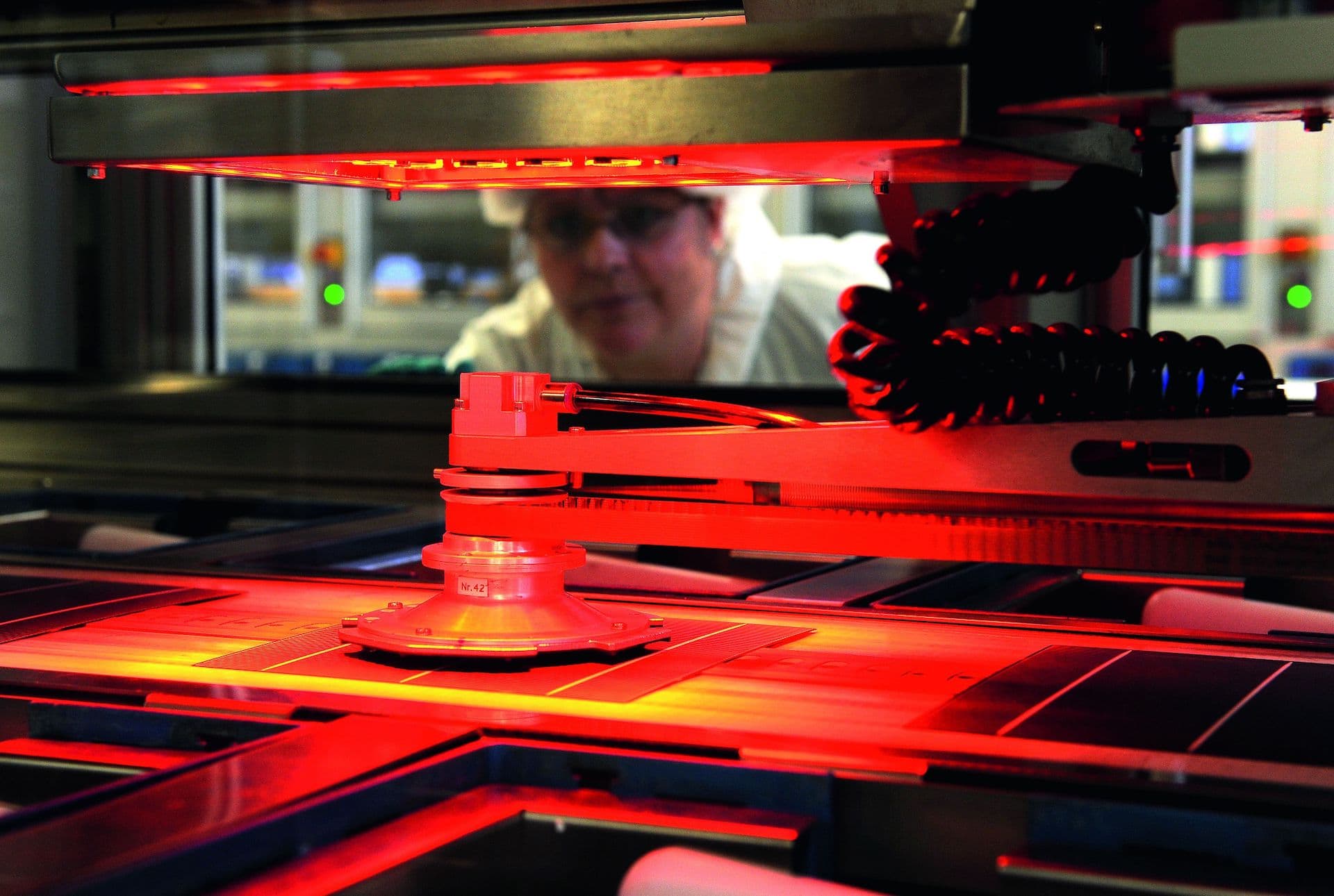 Advanced stage of silicon wafers within the solar module manufacturing process, under a red light with an engineer observing through a processing window to identify that silicon wafer prices have stabilised and and downstream effect to overall module costs is coming.